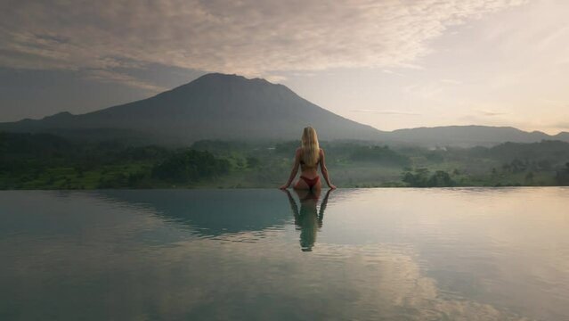 Fit woman in bikini on pool edge looking at magical sunrise with Volcano Agung