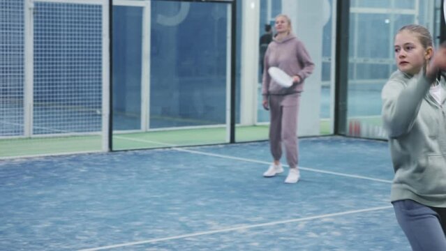 Teenage girl and adult woman playing padel in court against each other.