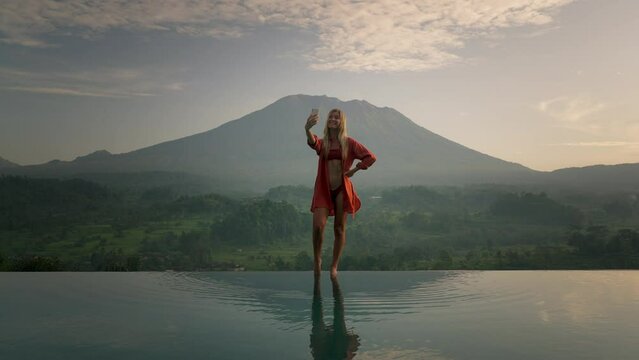 Blond model posing on infinity pool edge in bikini during sunrise, Bali Mount Agung
