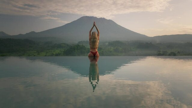 Woman in bikini with hourglass figure sitting on infinity pool edge yoga salute to Mount Agung