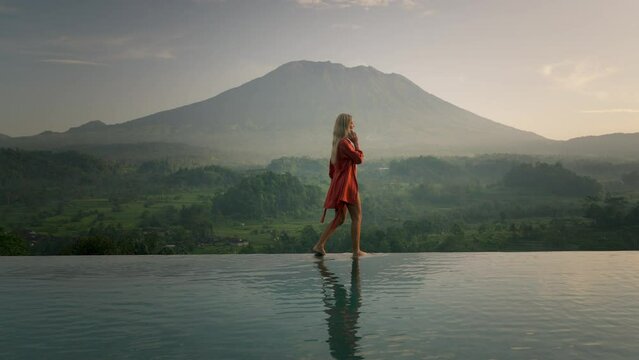 Blond business woman in silk bathrobe walking on infinity pool edge while on phone call, Mount Agung in background