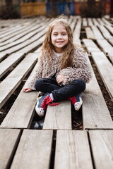 Portrait of little cute girl with long curly hair. Outdoor in the garden. Fashionable preschool child sitting on a wooden pallet in the park.