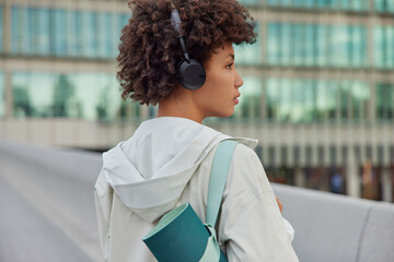Back view of curly haired woman listens music in headphones dressed in jacket carries rolled karemat focused away stands outdoors against blurred background. Healthy active lifestyle concept © WHstudio Leushin N