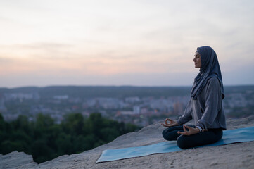 Beautiful young girl in a hijab meditates and practices yoga on the background of the cityscape at sunset