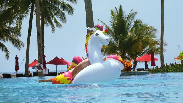 Happy funny man successfully jump onto floating inflatable unicorn in swimming pool in tropical hotel to ride. Smiling male tourist have fun on giant toy in water. Summer vacation, holiday concept.