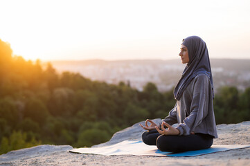 Beautiful young girl in a hijab meditates and practices yoga on a background of sunset and...