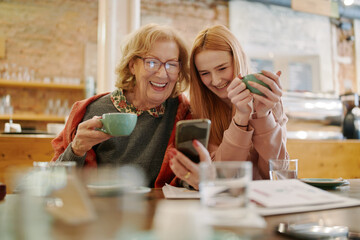 Happy grandmother and her adolescent granddaughter sitting in a cafe, enjoying coffee and using smart phone for funny videos. - 507778050