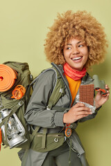 Vertical shot of happy female hiker holds bar of chocolate and binoculars smiles positively wears windbreaker carries rucksack has hike trip in mountains during summer hoidays leads active lifestyle © WHstudio Leushin N