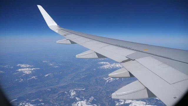 View from passenger cabin window of wing of aircraft flying over Austrian Alps on sunny day against background of blue sky and landscape with snow-capped rocky mountain ranges below. Travel concept 