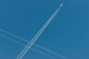 Samolot lecący po bezchmurnym, niebieskim niebie pozostawiający białą smugę kondensacyjną, krzyżującą się ze smugą przelatującego wcześniej samolotu.