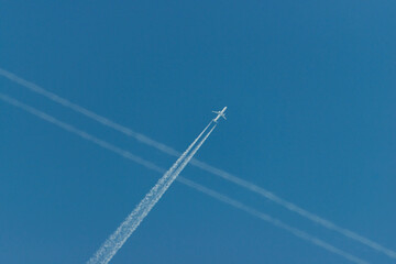 Samolot lecący po bezchmurnym, niebieskim niebie pozostawiający białą smugę kondensacyjną, krzyżującą się ze smugą przelatującego wcześniej samolotu. - obrazy, fototapety, plakaty