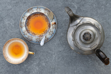 An old metal teapot, an old teacup with tea and saucer and an old teacup without saucer stand next...