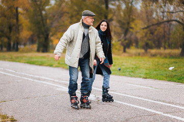 Funny, frightened happy pensioner in glasses ineptly roller-skates with his daughter in the autumn park. Active old age. - obrazy, fototapety, plakaty
