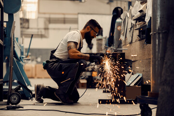 A heavy industry worker kneeling and grinding metal parts in factory.