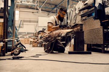 A heavy industry worker kneeling in facility and processing metal parts with grinder.