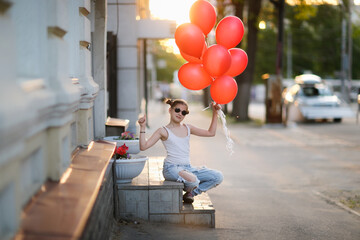 Funny stylish European girl child with red balloons in torn jeans sits on stairs on sunny summer...