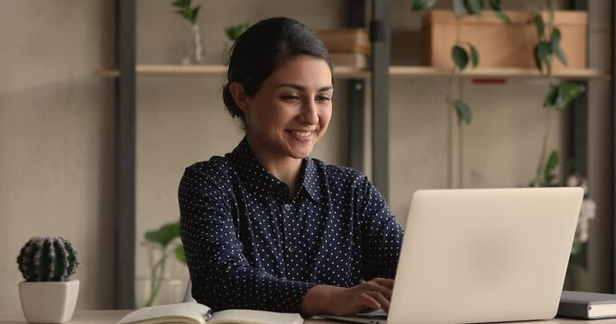 Indian woman sit at table in cozy office room working on laptop, freelancer texting e-mail response to client, make telework creative job from home using internet and modern wireless device concept