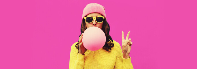 Fashionable portrait of stylish cool young woman inflating chewing gum wearing yellow knitted sweater, hat on pink background, blank copy space for advertising text