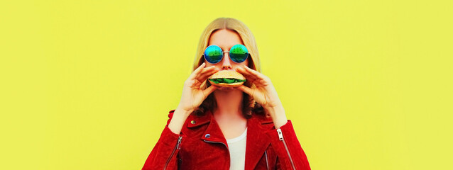Portrait of stylish happy smiling young woman with tasty big burger fast food on yellow background,...