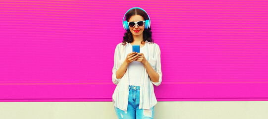 Portrait of happy smiling young woman in headphones listening to music with smartphone on pink background, blank copy space for advertising text