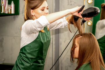 The hairdresser is drying long brown hair with a hairdryer and round brush in a beauty salon