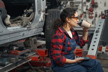 Woman car mechanic drinking coffee in automobile garage