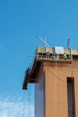 Using a construction gondola lift, workers place panels on a towering skyscraper. Building construction with blue sky as background