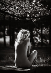 Naked woman sitting at a picnic table among pine trees.