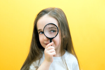 Positive curious schoolgirl in casual clothes looks at the camera through a magnifying glass, on a yellow background.