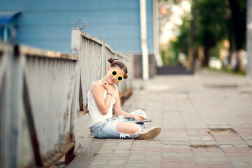 pretty girl with headphones and a phone sits on the sidewalk, a lonely child on a city street....