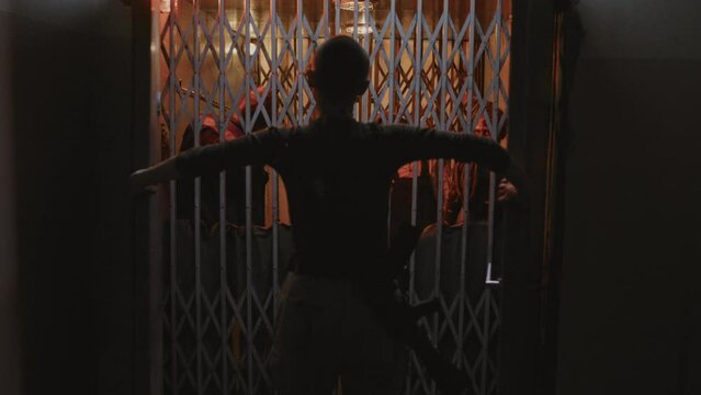Locked-down rear of military woman with shaved head walking in dark creepy building, opening doors, looking at three zombies stuck behind metal gate of old elevator reaching hands out