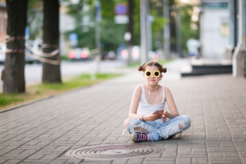 pretty girl with headphones and a phone sits on the sidewalk, a lonely child on a city street....