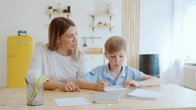 Young mother helping her son to do his homework at home, they sit at table in living room, communicating. Mom baby sitter nanny explaining to the boy task, school exercise, schoolboy writing.