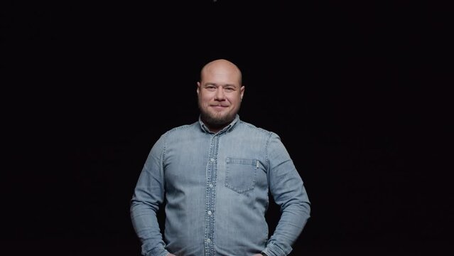A bald man with a beard in a denim shirt smiles and poses for the camera on a black background.