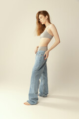 Full-length portrait of slim beautiful young woman posing in grey underwear and oversized jeans isolated over grey studio background
