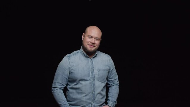 A bald man with a beard in a denim shirt looks in front and smiles. A man poses in front of the camera on a black background