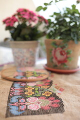 Traditional cross-stitched folk motif embroidery on natural burlap fabric background closeup on foreground with houseplants in flower pots on blurred background. Needlework embroidery crafting hobby