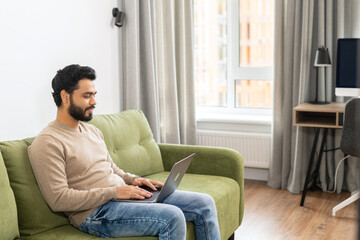 Positive Hindi man in smart casual clothes using computer while sitting at the sofa in his flat. Young Indian male student watching webinars, educational courses, learning on the distance