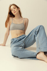 Portrait of young red-haired girl sitting on floor posing in oversized jeans and top isolated over grey studio background. Natural beauty