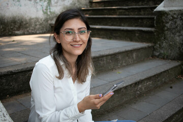 portrait of beautiful young girl smiling at the camera holding a smart phone on the stairs of a park