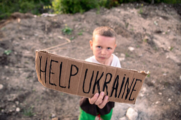 A boy with a grimy face and sad eyes holds a cardboard poster with the inscription HELP UKRAINE
