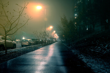 Street with poor visibility due to fog. Empty sidewalk illuminated by street lamps on a foggy night