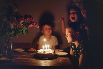 Cute child girl at table with birthday cake with siblings, dark style. Cake and candle 9 years birthday celebration