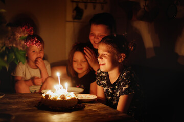 Cute child girl at table with birthday cake with siblings, dark style. Cake and candle 9 years birthday celebration
