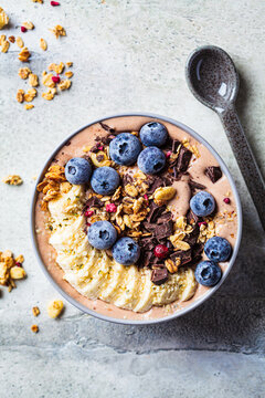 Chocolate banana smoothie bowl with granola, berries and hemp seeds, gray background. Healthy vegan recipe concept.