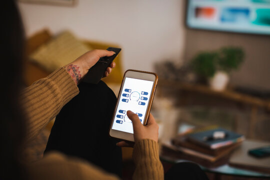 Cropped image of woman using app on smart phone while watching TV at home