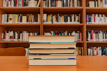 Pile of books on a table in a library room. In the background is a bookshelf that houses the...