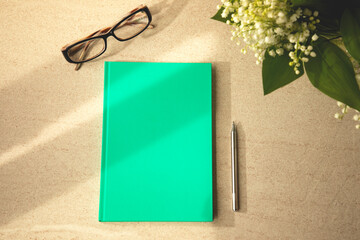 Green notebook on a brown table with a pen, glasses and a bouquet of lilies of the valley. Office table and stationery with window shadow, flat lay