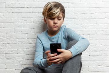 A schoolboy sits with a phone in his hands against the background of a white brick wall. A handsome guy in a blue sweater and gray pants. Chats, games and social networks.