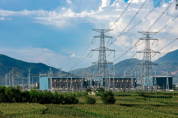 High voltage towers and substations stand on a corn field under a blue sky and white clouds.
